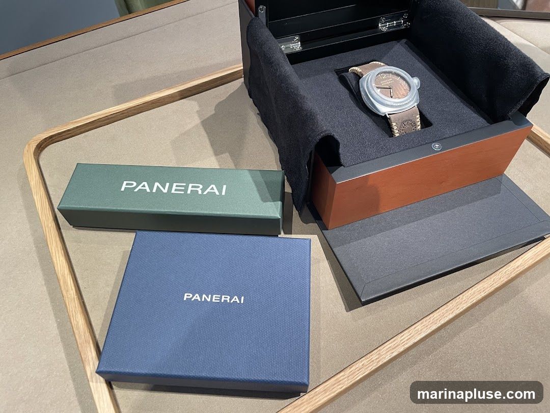 A Panerai watch is displayed in its open wooden and black velvet box. Next to the watch box are two smaller Panerai branded boxes, one green and one blue. The items are arranged on a textured surface with a wooden border.