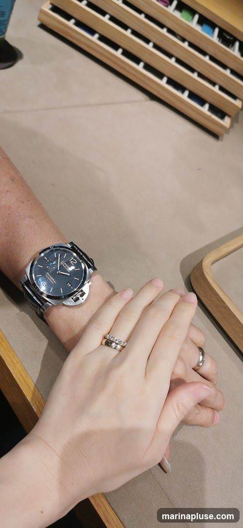 A close-up shot of two people's hands, one wearing a large wristwatch and both wearing rings. The background shows a display case with multiple compartments, possibly for jewelry or accessories.