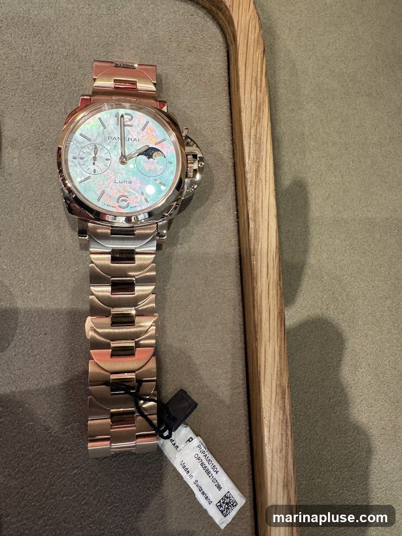 A rose gold Panerai watch with a mother-of-pearl dial featuring a moon phase complication. The watch has a date window and a small seconds subdial. A tag attached to the bracelet reads "PNPAM01504 OP7605BB2107266 Made in Switzerland". The watch is displayed on a textured surface next to a wooden stand.