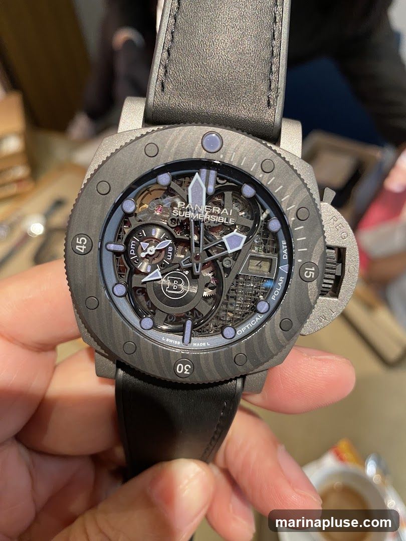 A close-up shot of a Panerai Submersible watch with a black leather strap. The watch face is skeletonized, revealing the intricate inner workings. The dial features luminous hands and markers, with "PANERAI SUBMERSIBLE" clearly visible. The bezel has markings for minutes and a date window. The watch is held by a person, with a blurred background suggesting an indoor setting.