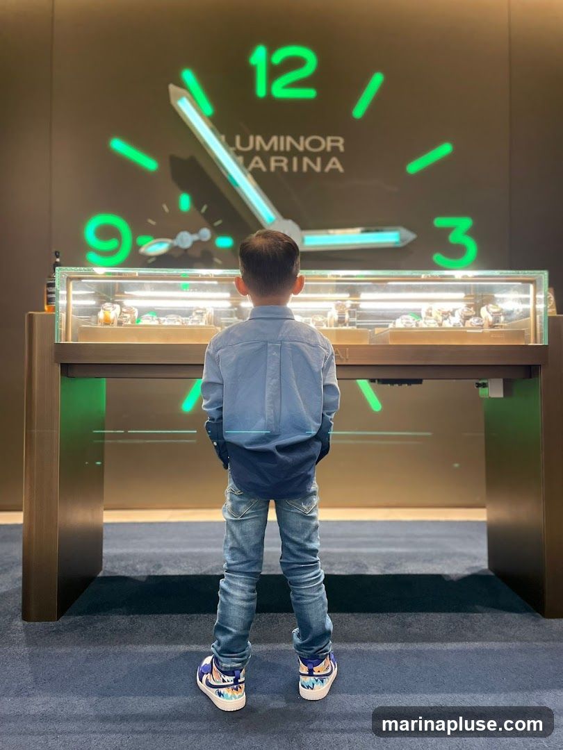 A young boy stands with his back to the camera, looking at a display case filled with watches. Behind him, a large clock face with the words "LUMINOR MARINA" is illuminated in green neon. The display case is lit from within, showcasing various timepieces. The floor is carpeted in dark blue, and the walls are a dark brown.