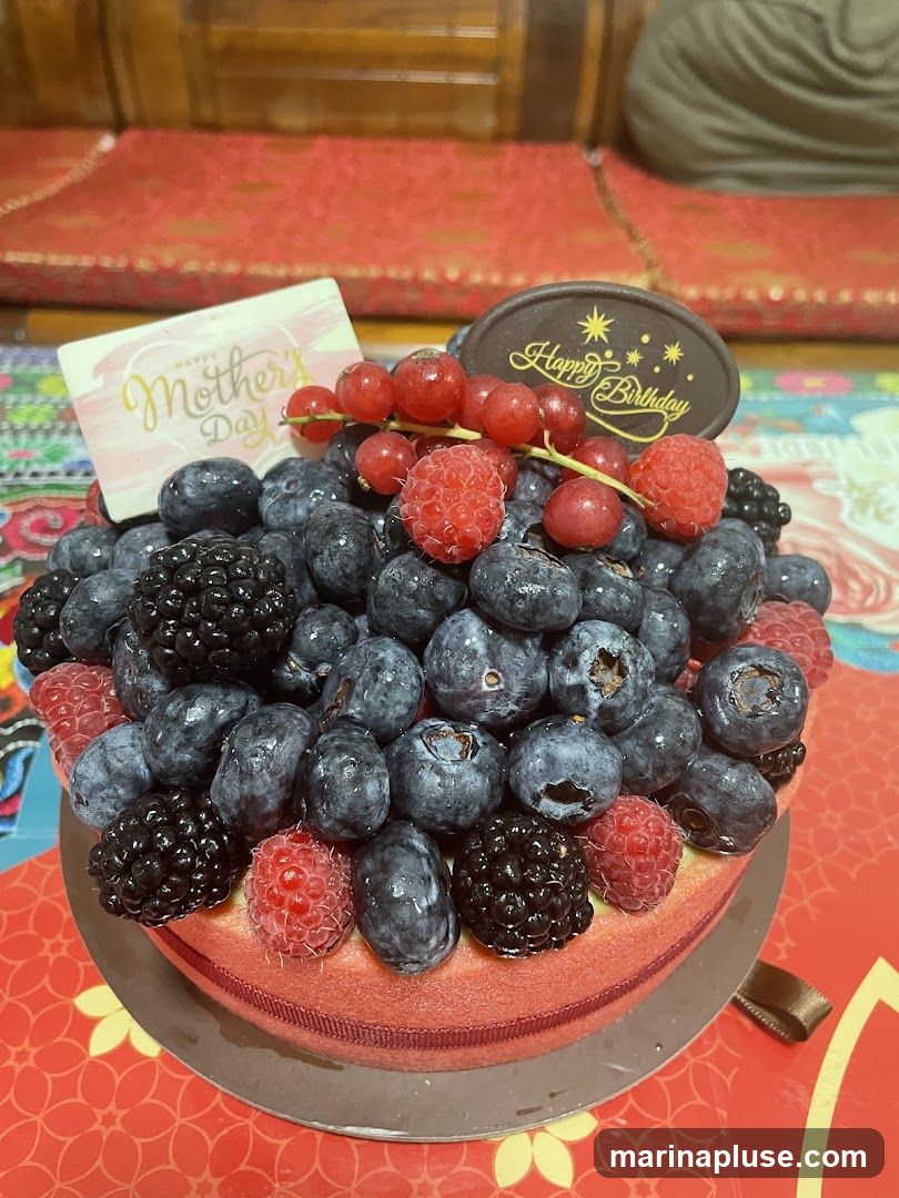 A round cake topped with blueberries, raspberries, blackberries, and red currants. A white card with "Happy Mother's Day" in gold lettering and a chocolate oval with "Happy Birthday" are placed on top. The cake is on a decorative surface.