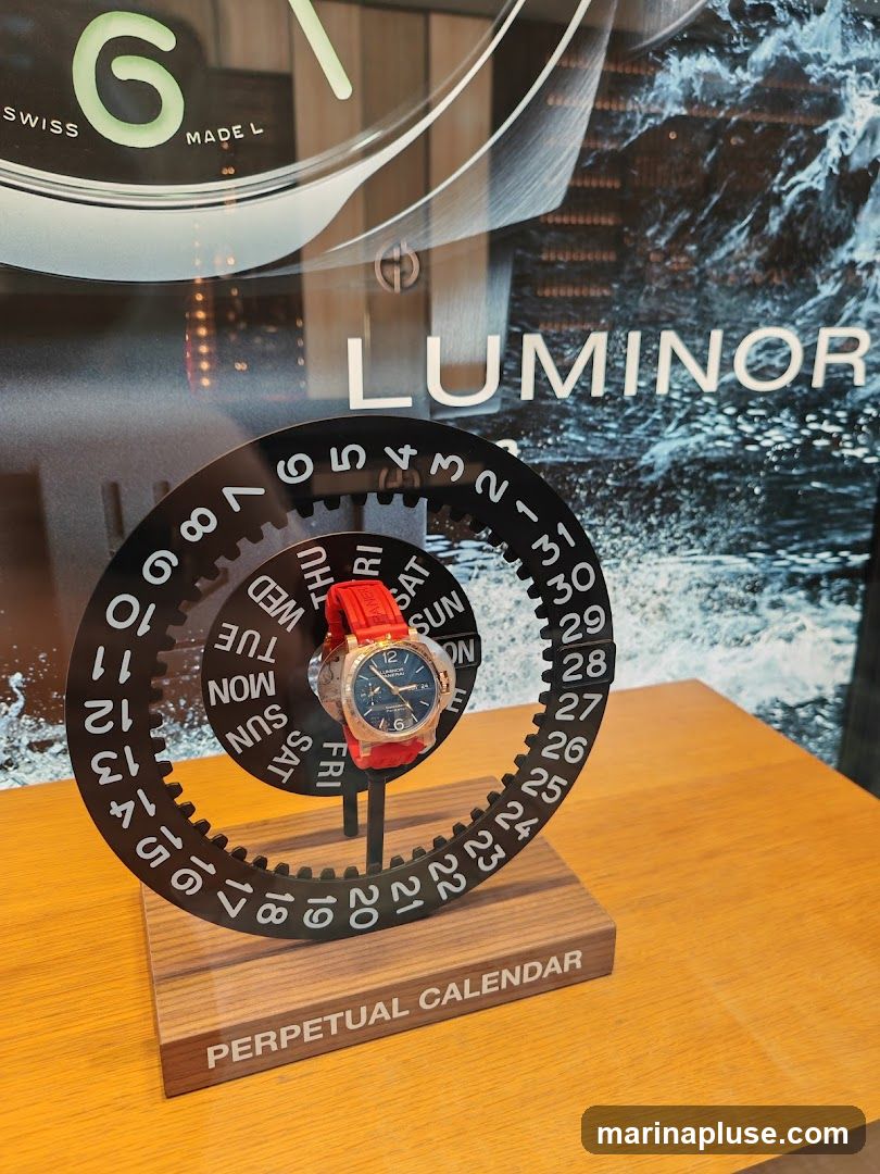 A close-up view of a watch display featuring a watch with a red strap. The watch is placed on a stand with a circular calendar mechanism. The background shows a large advertisement with the word "LUMINOR" and a water-themed design. The text "PERPETUAL CALENDAR" is visible on the base of the stand.