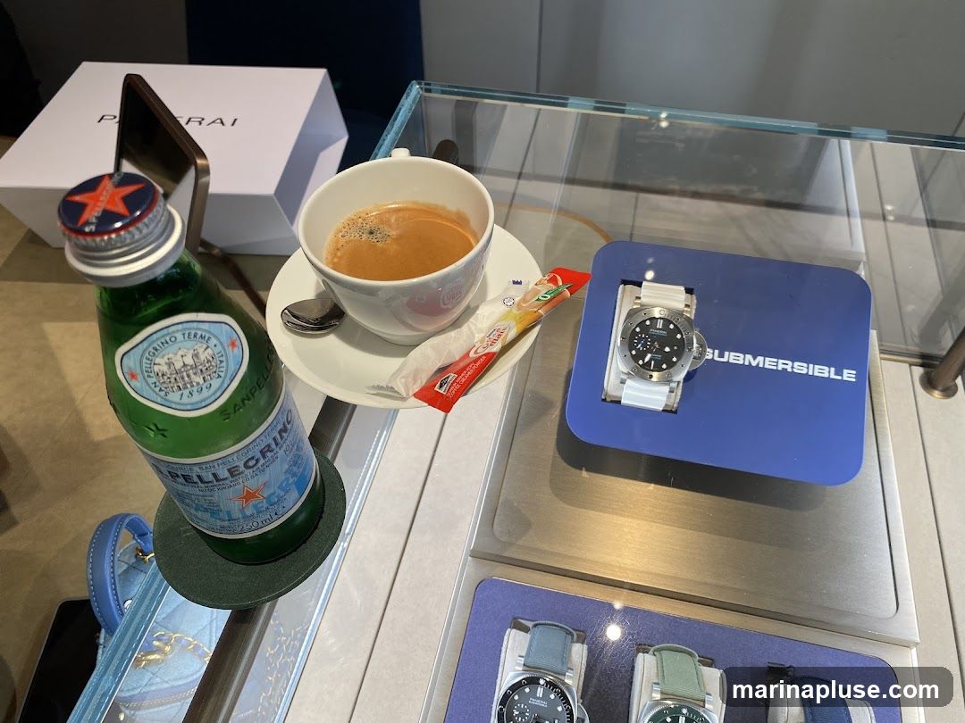 A close-up view of a display case featuring a watch on a blue stand with the word "SUBMERSIBLE" visible. Next to it, a cup of coffee sits on a saucer with a sugar packet. A bottle of San Pellegrino water is also present, along with a Panerai box in the background. The display appears to be in a retail environment, possibly a watch store.