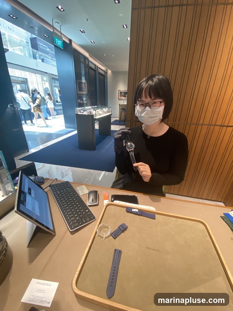 A woman wearing a mask and gloves holds a watch in a brightly lit watch store. In front of her is a display tray with watch straps and a watch case. A laptop, keyboard, and mouse are on the counter. The background shows display cases with watches and an exit sign.