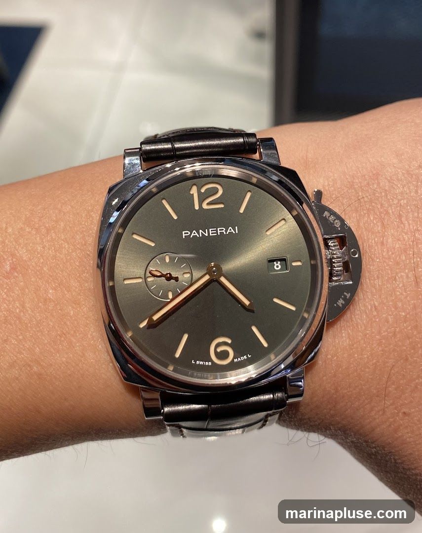 A close-up shot of a Panerai watch on a person's wrist. The watch has a dark grey dial with luminous hour markers and hands. The number 12 is prominent at the top, and the number 6 is at the bottom. A small sub-dial for seconds is visible at the 9 o'clock position, and a date window is at the 3 o'clock position. The watch features a polished metal case and a dark leather strap. The crown guard is a distinctive feature on the right side of the case.