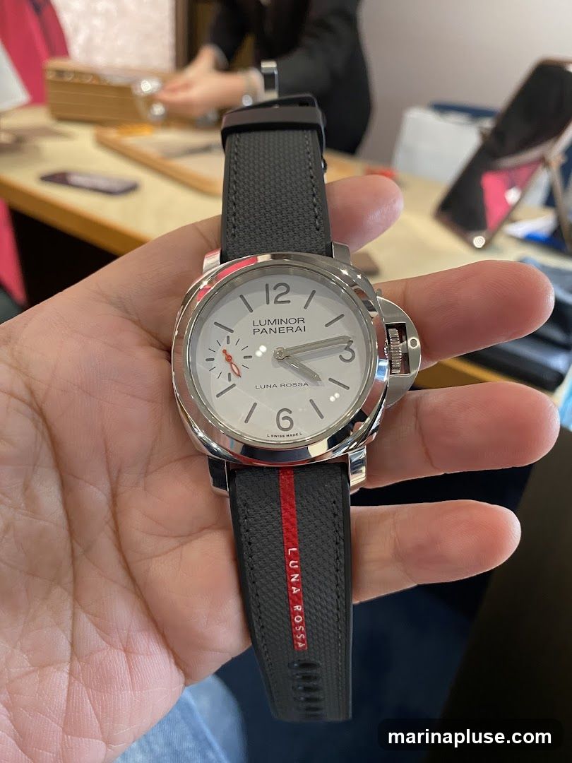 A close-up shot of a Panerai Luminor Luna Rossa watch held in a person's hand. The watch has a white dial with silver hour markers and hands. The strap is dark gray with a red stripe and the text "LUNA ROSSA" printed in red. In the blurred background, a person appears to be working at a counter in a store.
