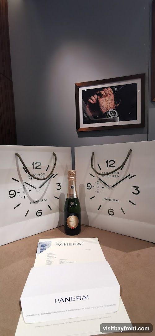 A display featuring two Panerai shopping bags designed to look like watch faces, a bottle of champagne, and Panerai branded paperwork. A framed picture of a watch on a wrist hangs on the wall above.