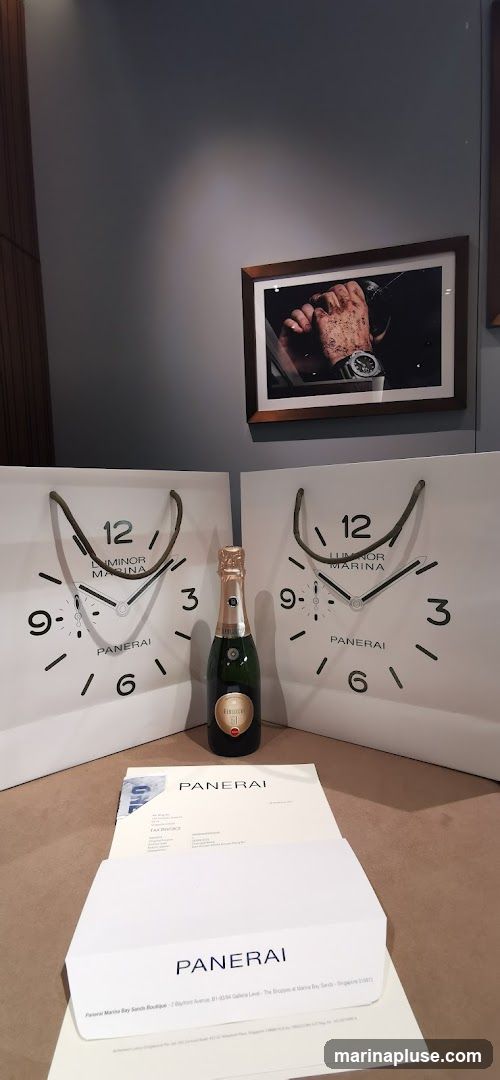 A display featuring two Panerai shopping bags designed to look like watch faces, a bottle of champagne, and Panerai branded paperwork. A framed picture of a watch on a wrist hangs on the wall above.