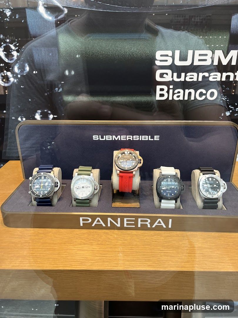 A display case featuring five Panerai Submersible watches. The watches have various strap colors including blue, green, red, white, and black. The display is set against a backdrop with text that reads "SUBM" and "Quarant" and "Bianco". The brand name "PANERAI" is prominently displayed below the watches.