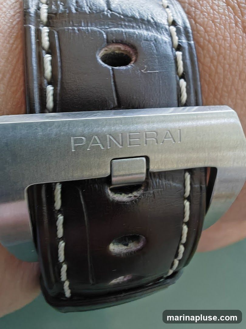 A close-up image of a Panerai watch clasp and dark brown leather strap with white stitching. The clasp is made of brushed metal and has the word "PANERAI" engraved on it.