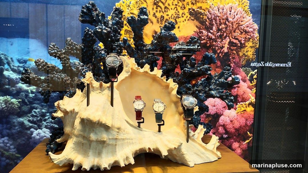 A display featuring several watches presented within a large seashell. The backdrop is a vibrant underwater scene with colorful coral and fish. The watches are arranged on stands, with some featuring dark straps and others with red or grey straps. The overall presentation evokes an oceanic theme.