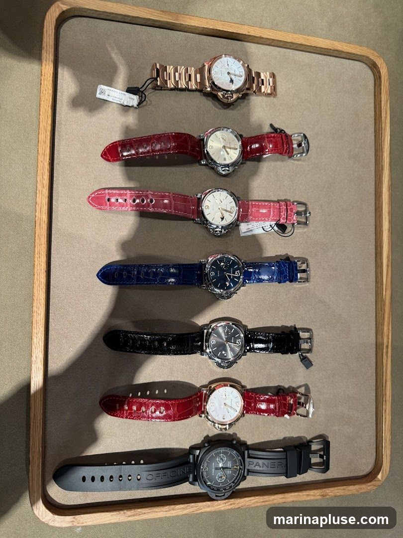 A display of seven watches with various strap colors including rose gold, red, pink, blue, black, and red. The watches are laid out horizontally on a neutral-colored surface within a wooden tray. Some watches have visible brand names like "OFFICINE PANERAI" on their straps.