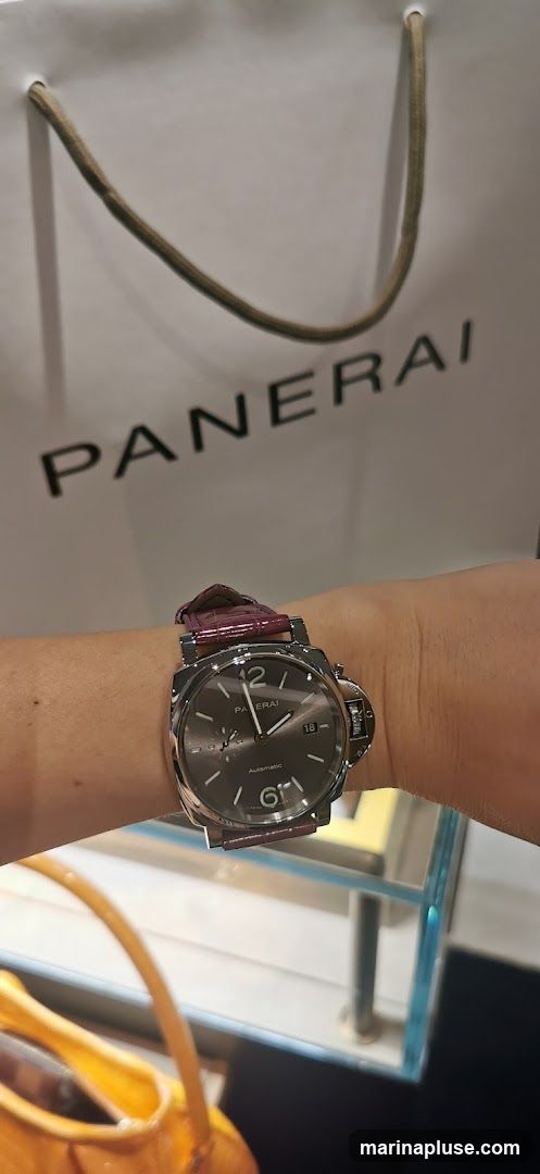 A close-up shot of a person's wrist wearing a Panerai watch with a dark dial and a burgundy strap. In the background, a white shopping bag with the word "PANERAI" printed in large black letters is visible. A yellow handbag is partially visible in the foreground. The scene appears to be inside a store, with a glass display case in the lower right corner.