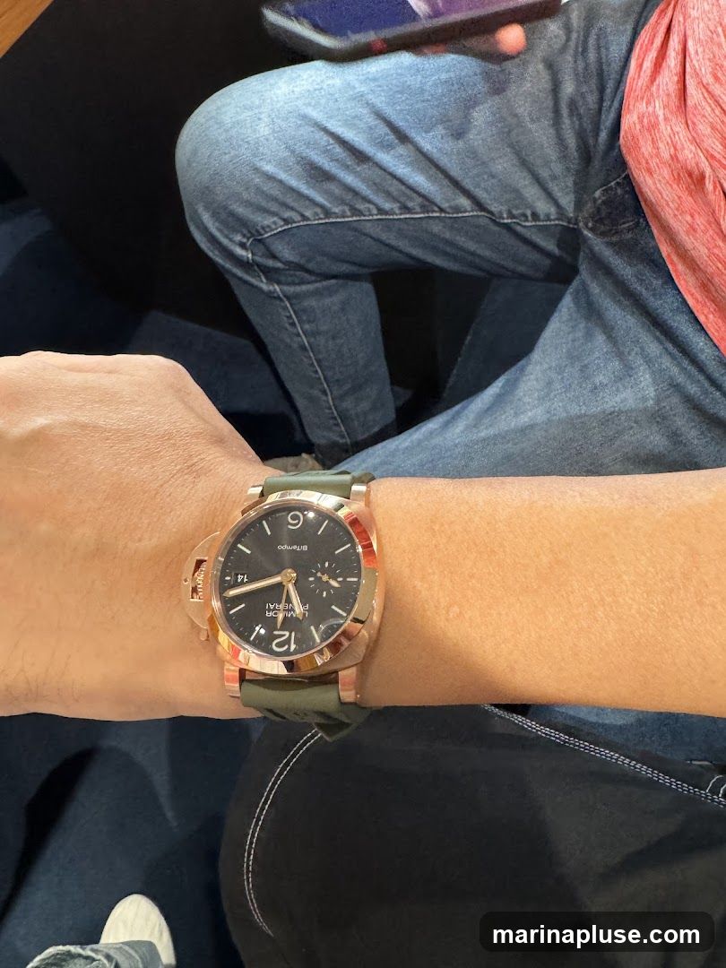 A close-up shot of a person's wrist wearing a rose gold watch with a dark dial and a green strap. The watch face shows Arabic numerals and has a small sub-dial. The person is wearing blue jeans and a red shirt.