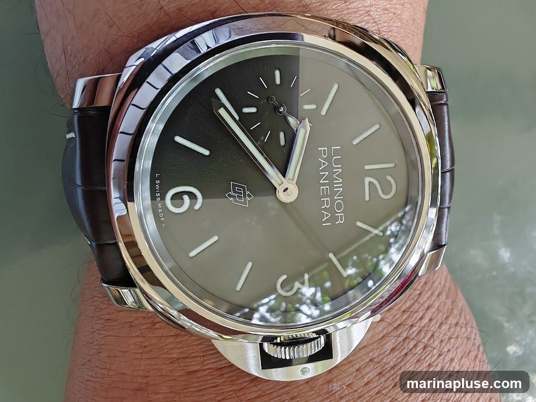 A close-up shot of a Panerai Luminor watch on a wrist. The watch has a dark grey dial with white hour markers and hands. The brand name "PANERAI" and model "LUMINOR" are visible on the dial. The watch has a brown leather strap and a polished stainless steel case. The crown guard is also visible.
