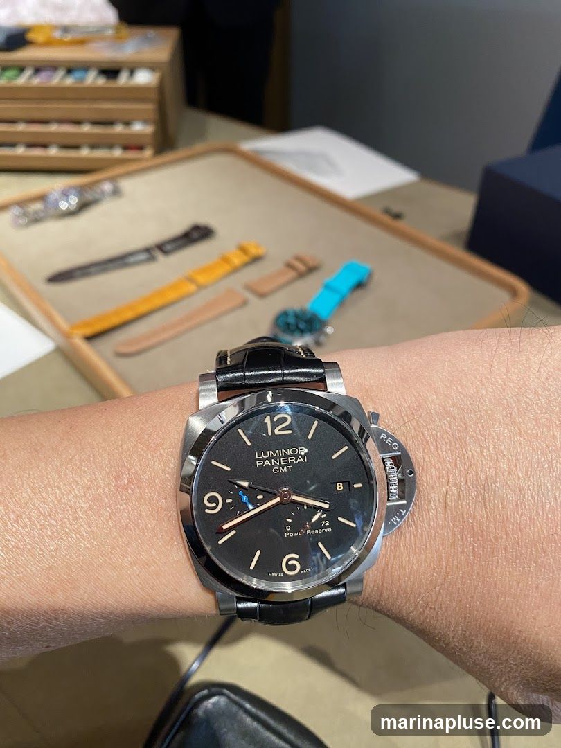 A close-up shot of a Panerai Luminor GMT watch on a person's wrist. The watch has a black dial with luminous hour markers and hands, a date window at 3 o'clock, and a power reserve indicator. The watch is on a black leather strap. In the background, there is a display tray with various watch straps and a wooden organizer with compartments.