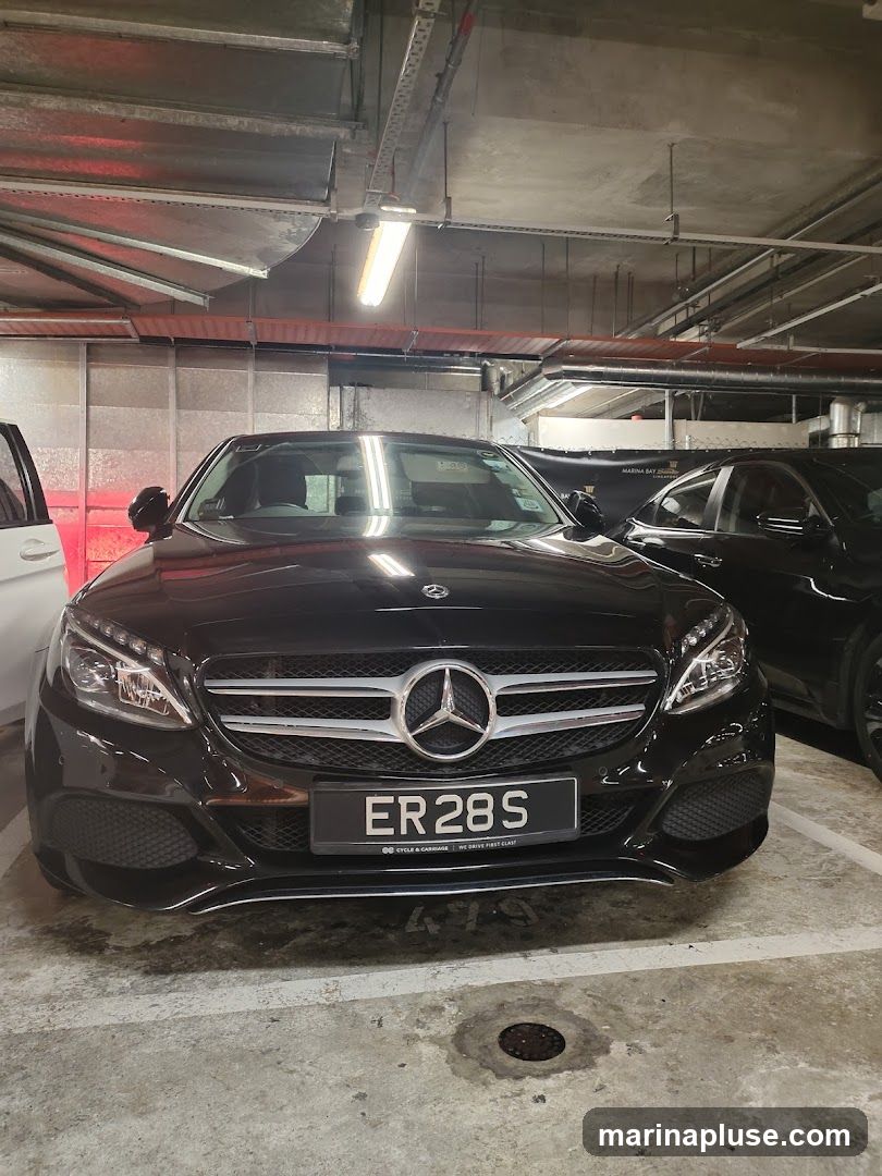 A black Mercedes-Benz C-Class is parked in a dimly lit parking garage. The car's license plate reads "ER 28 S". The car is facing the camera, and its headlights are on. The background shows other cars and the industrial ceiling of the parking garage with exposed pipes and lighting fixtures.