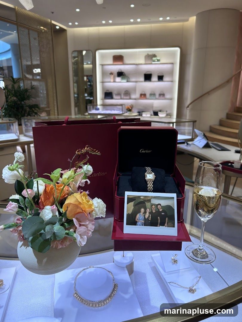 A Cartier display features a watch in an open red box, a bouquet of flowers, a glass of champagne, and several pieces of jewelry on display. A photograph of three people is placed in front of the watch box. In the background, shelves are stocked with handbags.