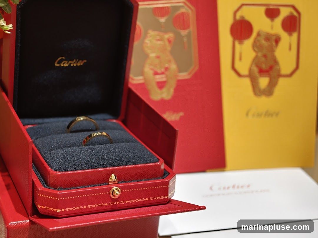 A red Cartier ring box is open, displaying two gold rings inside. Behind the box are red and yellow gift bags with a bear and lantern design, also branded with Cartier. A white Cartier envelope is in the foreground.