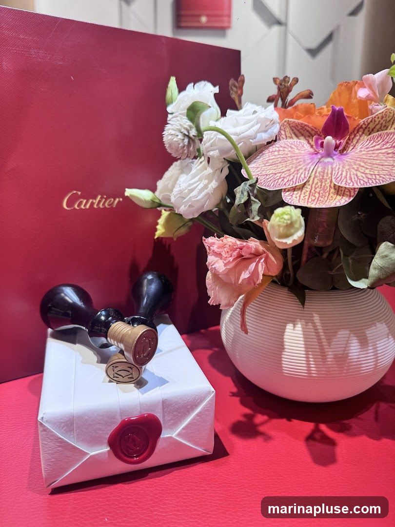 A red Cartier shopping bag is partially visible on the left. In front of it, a white gift-wrapped box is adorned with a red wax seal featuring the Cartier logo. Two stamps, also with the Cartier logo, rest on top of the gift. A vase of colorful flowers, including orchids and roses, is placed to the right of the gift. The items are arranged on a red surface.