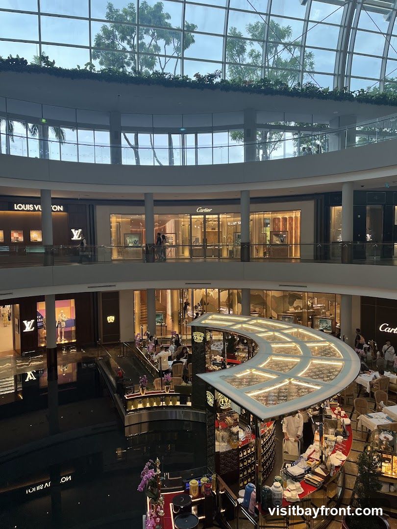 The image shows the interior of a multi-level shopping mall with a glass ceiling and a canal running through the center. Several luxury brand stores, including Louis Vuitton and Cartier, are visible on the upper levels. A curved bar or cafe area is situated over the canal, with patrons seated at tables. The overall ambiance is upscale and modern.