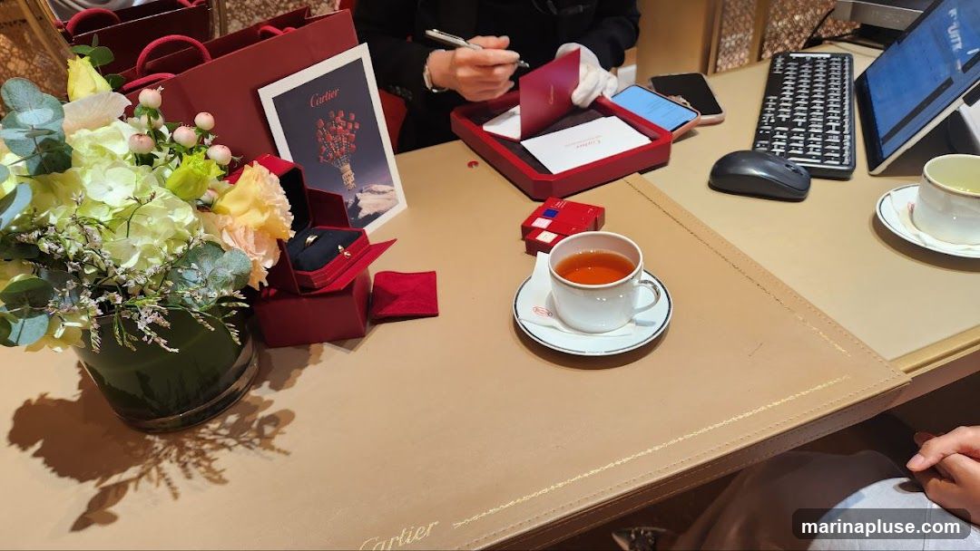 A table at Cartier with a bouquet of flowers, jewelry boxes, a cup of tea, and a person signing a card. There are also shopping bags and electronic devices on the table.