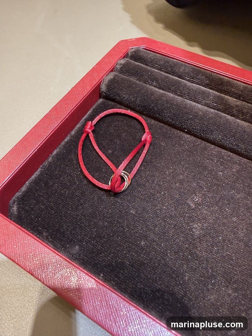 A red cord bracelet with three interlocking rings is displayed in a red jewelry box with a black velvet lining. The bracelet is tied with a knot.