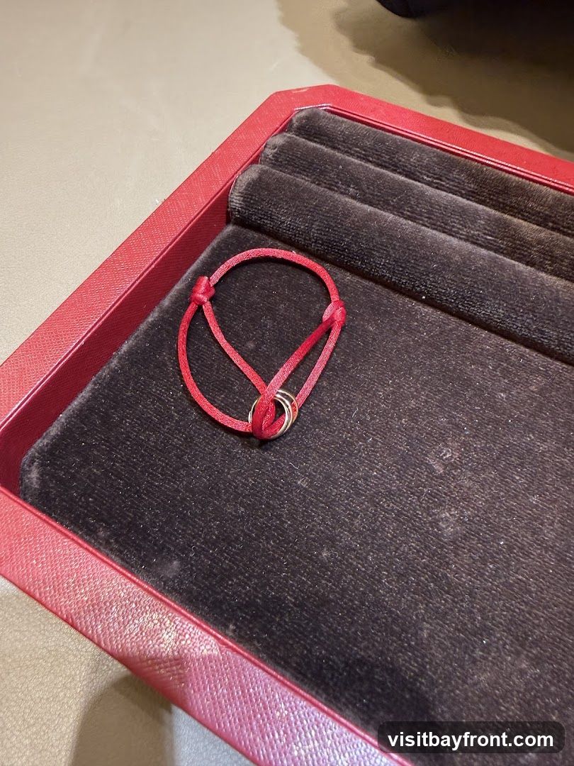 A red cord bracelet with three interlocking rings is displayed in a red jewelry box with a black velvet lining. The bracelet is tied with a knot.