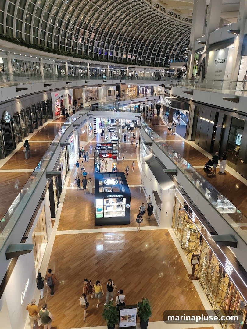 The image shows a wide view of the interior of a multi-level shopping mall with a distinctive arched glass roof. The floors are made of polished wood, and there are numerous shops and escalators visible. People are seen walking around and browsing. In the center, there is a display kiosk for Montblanc. On the right, a store with a mirrored facade is visible, and further down, a store with the name 'BIRKENSTOCK' is displayed.