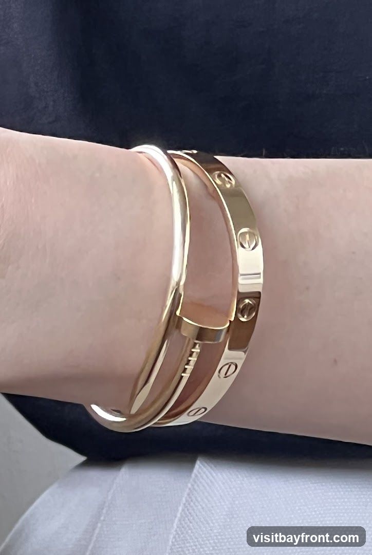 A close-up shot shows two gold bracelets on a person's wrist. The bracelet on top is a thin, smooth band. The bracelet underneath is wider and features engraved oval shapes with a line through them, resembling screws. The person is wearing a dark top and white pants.