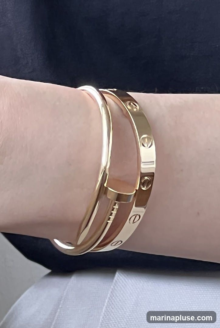 A close-up shot shows two gold bracelets on a person's wrist. The bracelet on top is a thin, smooth band. The bracelet underneath is wider and features engraved oval shapes with a line through them, resembling screws. The person is wearing a dark top and white pants.
