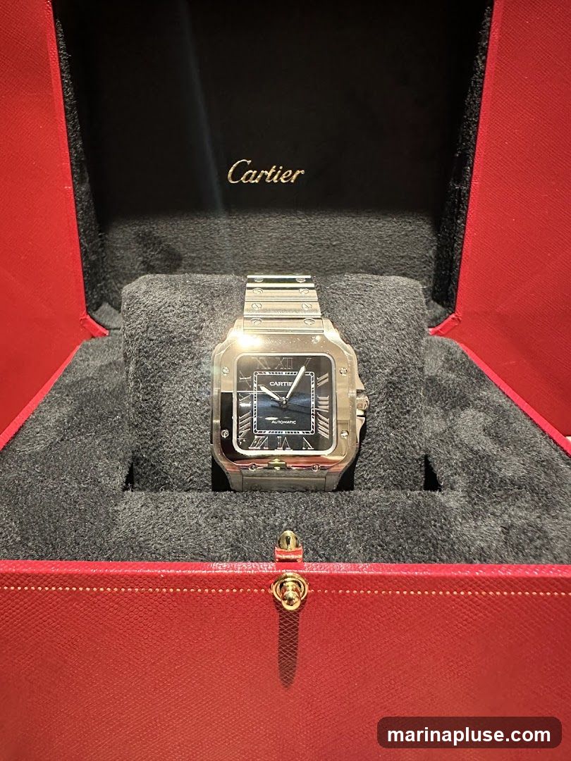 A silver Cartier watch with a black face is displayed inside a red and black Cartier presentation box. The Cartier logo is visible in gold lettering on the inside lid of the box.