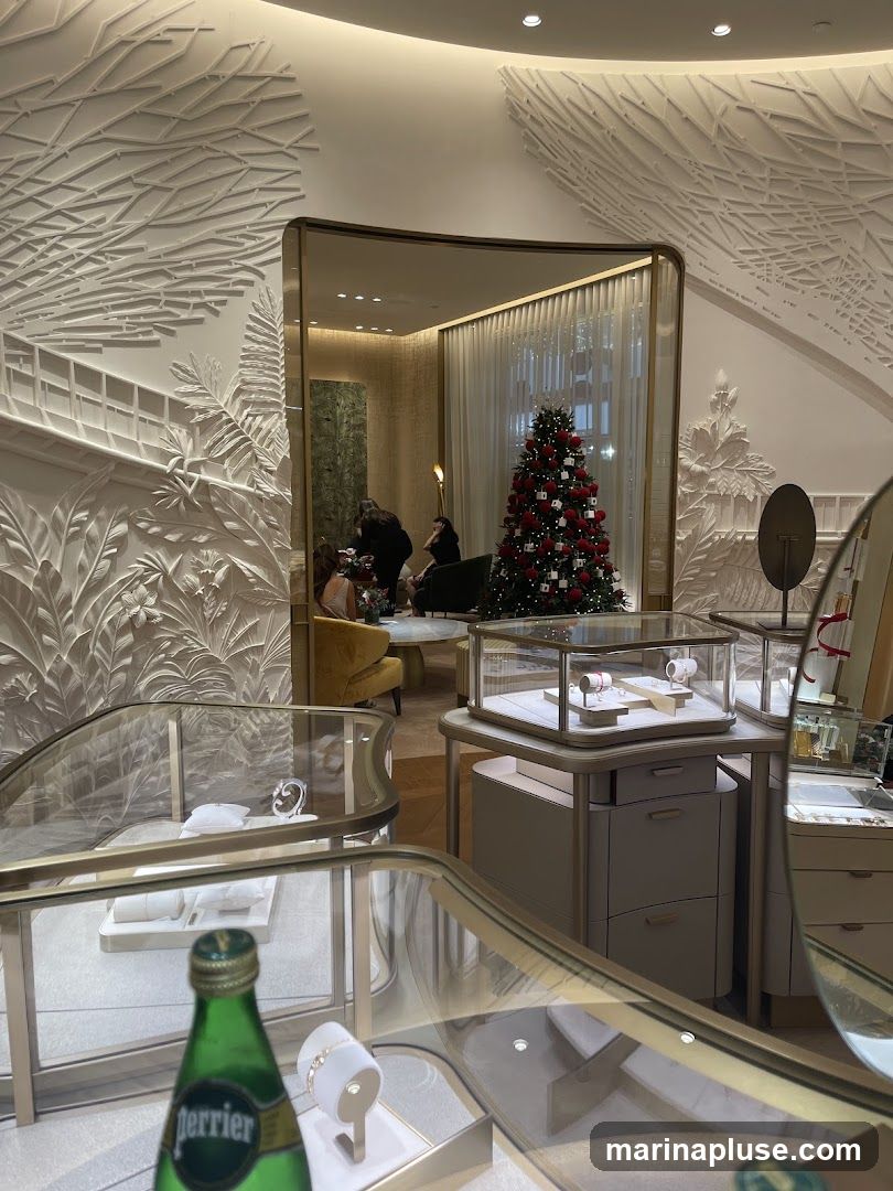 The interior of a high-end jewelry store is decorated for the holidays with a Christmas tree. Display cases showcase watches and jewelry. In the background, a seating area with plush chairs is visible, and several people are seated. The walls feature intricate, textured white molding resembling branches and leaves. A bottle of Perrier water is in the foreground.