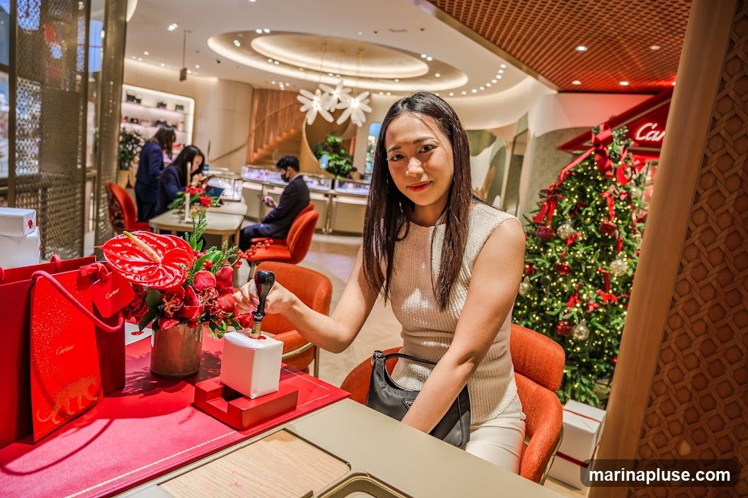 A woman is seated at a table in a brightly lit, upscale store, stamping a small white gift box with a black seal. Red Cartier shopping bags and a floral arrangement are on the table. In the background, other customers are browsing displays, and a decorated Christmas tree is visible. The store interior features elegant lighting and displays.