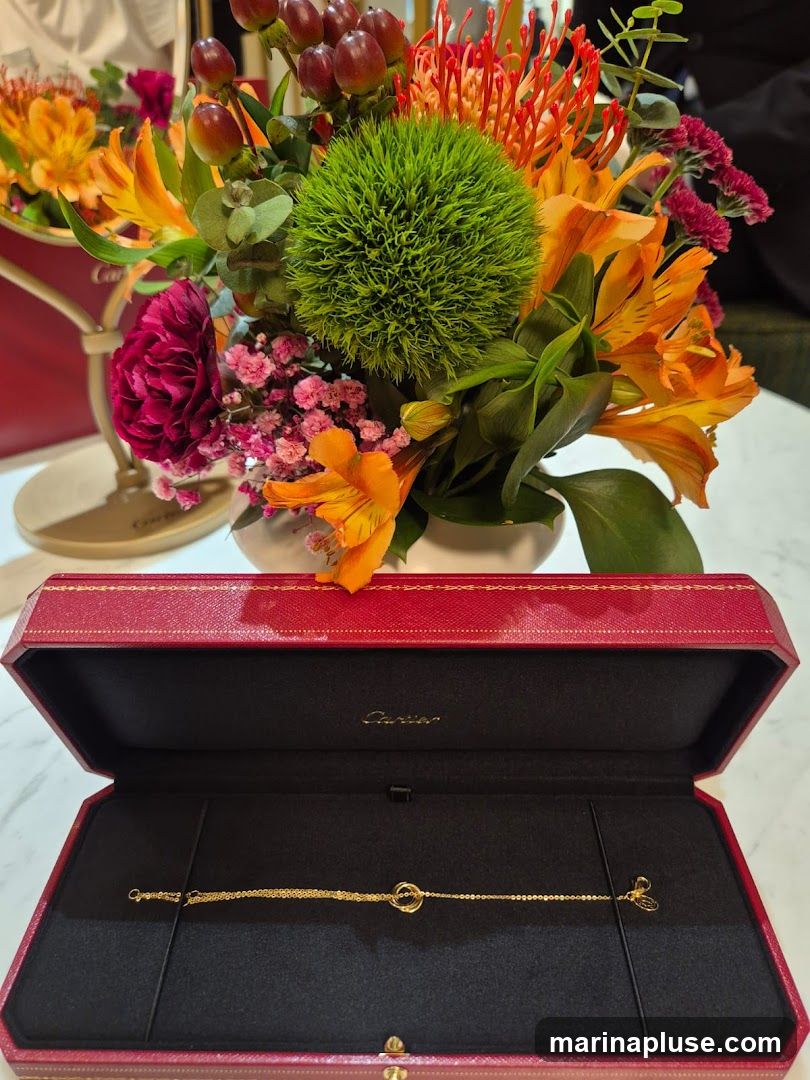 A gold bracelet with interlocking rings is displayed in an open red Cartier jewelry box. The box is placed on a white marble surface. Behind the box, a vibrant bouquet of flowers in shades of orange, red, pink, and green is arranged in a vase. The Cartier logo is visible inside the box lid.