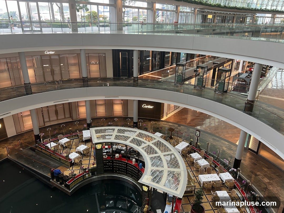 An aerial view of the interior of a modern shopping mall. On the upper levels, several storefronts are visible, including Cartier and Montblanc. Display cases with merchandise are placed along the walkways. On the lower level, a circular cafe or restaurant area is set up with tables and chairs around a central bar. The mall features a water feature or canal in the foreground.