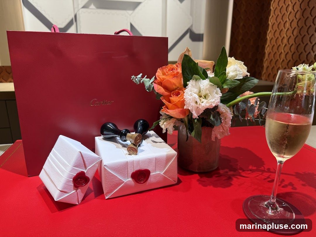A red Cartier shopping bag is displayed behind two white gift-wrapped boxes, each sealed with red wax. A pair of black and gold cufflinks with the Cartier logo are placed on top of one of the boxes. A vase of orange and white flowers sits to the right of the gifts, and a glass of champagne rests on a red coaster. The items are arranged on a red surface, likely a table.