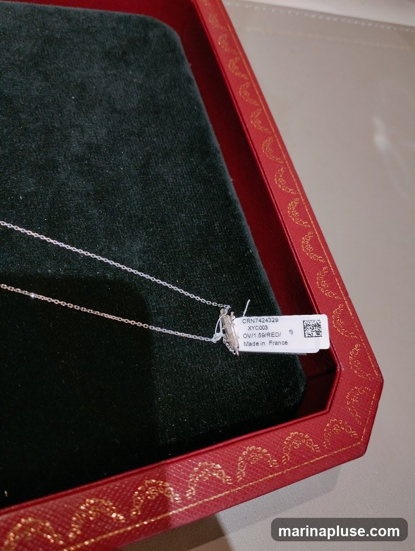 A close-up shot of a delicate diamond necklace resting on a dark green velvet surface within a red, gold-trimmed box. A white tag attached to the necklace displays product information including a reference number, code, material description, and origin.