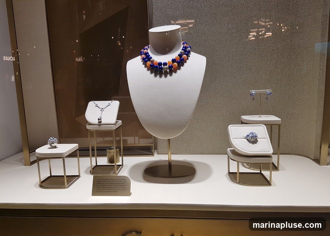 A display window showcases various pieces of jewelry, including necklaces, rings, and earrings, arranged on white stands and mannequins. The background is a textured wall with reflections of lights. A small plaque with text is visible on the lower left.