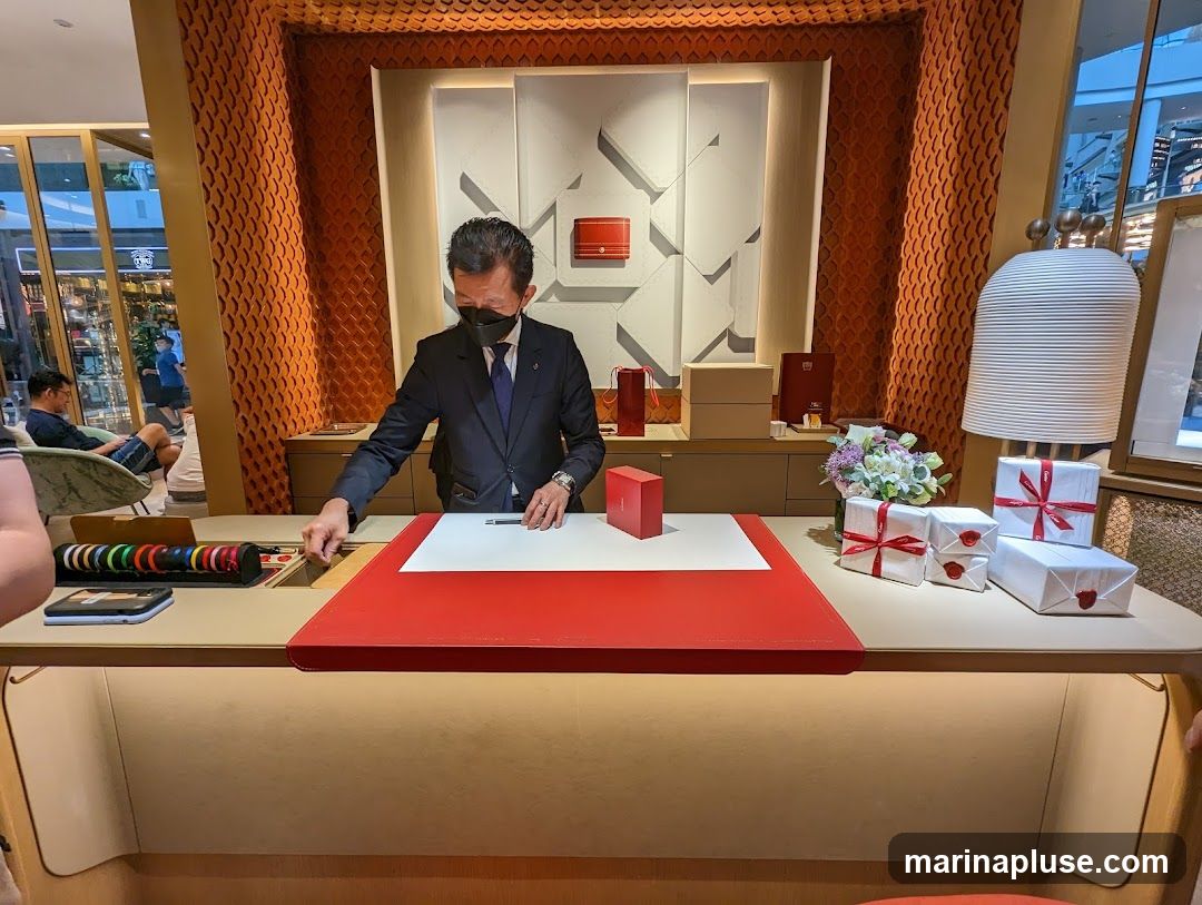 A sales associate in a suit and mask stands behind a counter, reaching into a drawer. The counter has a red surface and is adorned with wrapped gifts tied with red ribbon, a bouquet of flowers, and various boxes. The background features textured orange walls and a display with geometric patterns. In the background, through a glass partition, a shopping mall interior is visible with other shoppers and displays.