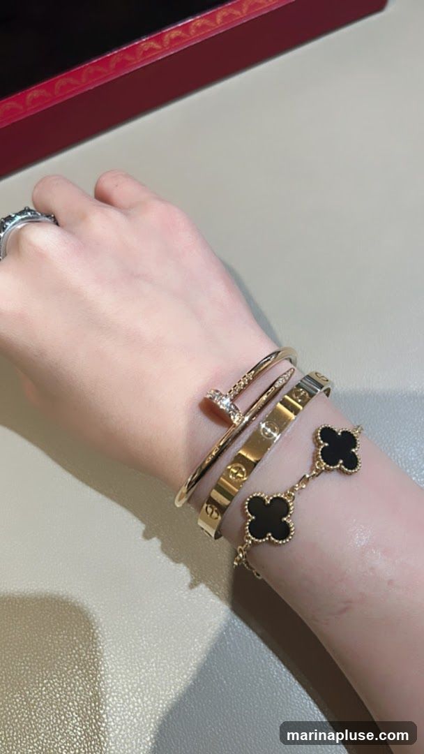 A person's wrist is adorned with three stacked bracelets. The top bracelet is a thin gold band with a diamond-encrusted nail head design. Below it is a wider gold bracelet with engraved logos. The bottom bracelet is a delicate gold chain featuring black clover-shaped charms. A red and gold box is visible in the upper left corner.