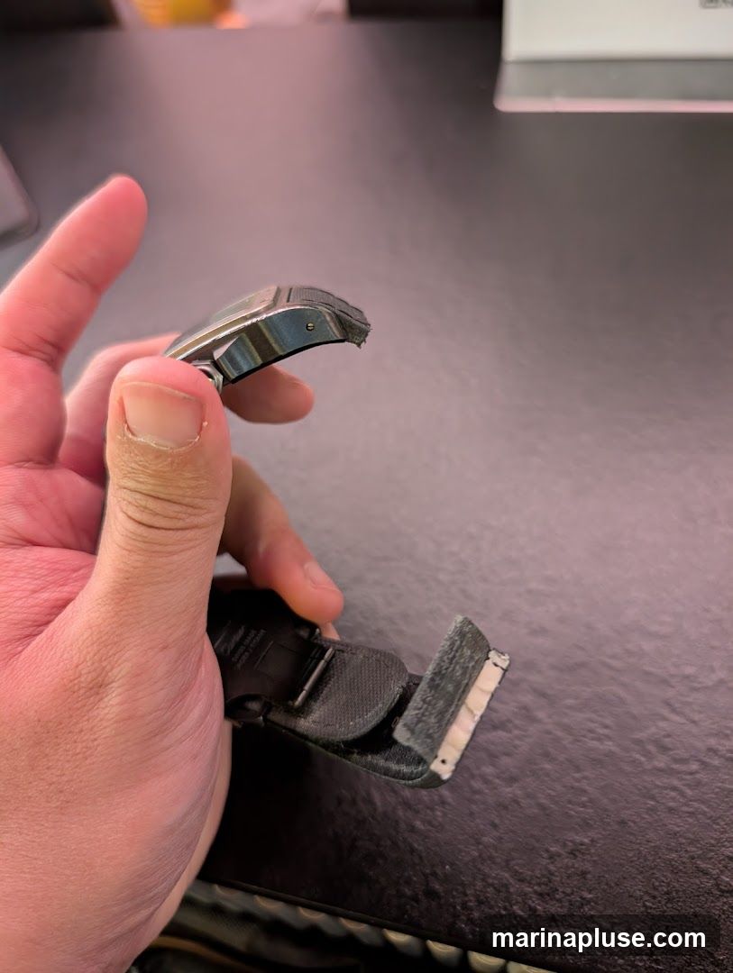 A person's hand holds a broken watch band. The band appears to be made of black plastic or rubber and is detached from the watch face. The material is frayed and damaged at the point of separation.