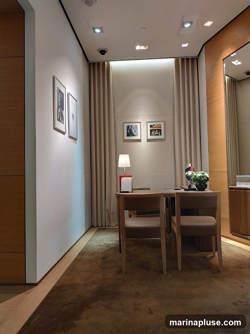 The image shows a well-lit, elegantly decorated interior of what appears to be a luxury boutique. A wooden desk with two chairs is centrally placed, with a lamp, a small floral arrangement, and a red and white display stand on its surface. The walls are a neutral tone, adorned with framed pictures. Beige curtains hang in the background, and a large mirror is visible on the right side of the frame. The floor is covered with a plush brown carpet.