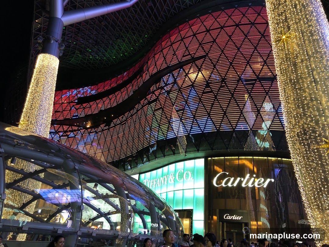 A nighttime view of the exterior of a building featuring storefronts for Cartier and Tiffany & Co. The architecture is modern and illuminated with colorful lights. A large, curved structure covered in lights is on the right, and another illuminated column is on the left. The facade of the building has a distinctive diamond-patterned grid with colored lights.