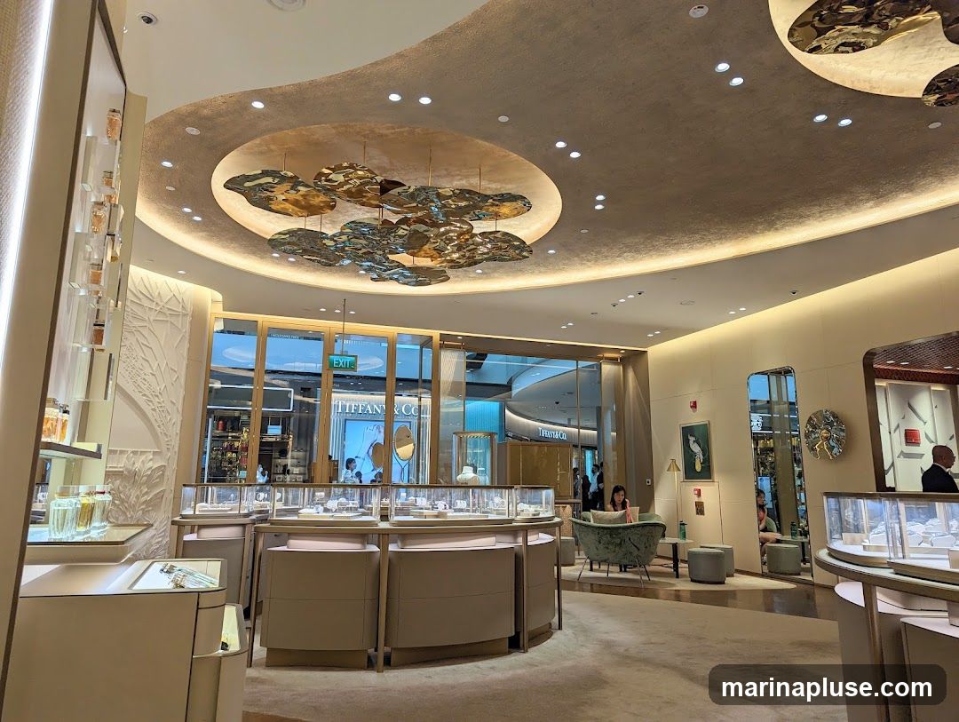 The interior of a high-end jewelry store with display cases filled with merchandise. The store features a modern and elegant design with a textured ceiling, decorative lighting, and comfortable seating areas. Through the glass doors, another store with the Tiffany & Co. logo is visible.