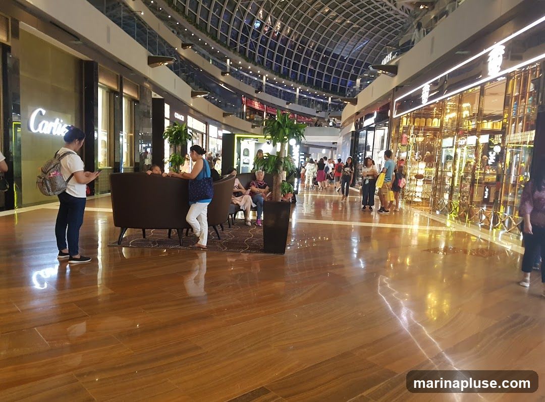 A wide shot of a modern shopping mall interior with a high, arched glass ceiling. The polished wooden floor reflects the bright lights and the storefronts. Several people are browsing or walking through the mall. On the left, a store with a prominent "Cartier" sign is visible. Further down, a store with a gold-toned, reflective facade displays various items. Seating areas with dark sofas are placed in the walkway, with some shoppers resting. Green potted plants are strategically placed throughout the mall.