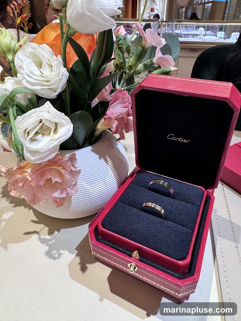A red Cartier ring box is open, displaying two gold rings with three horizontal bands. The box is placed next to a white vase filled with a colorful arrangement of flowers, including white roses, pink carnations, and orange blossoms. The background shows a blurred interior of a jewelry store with display cases.