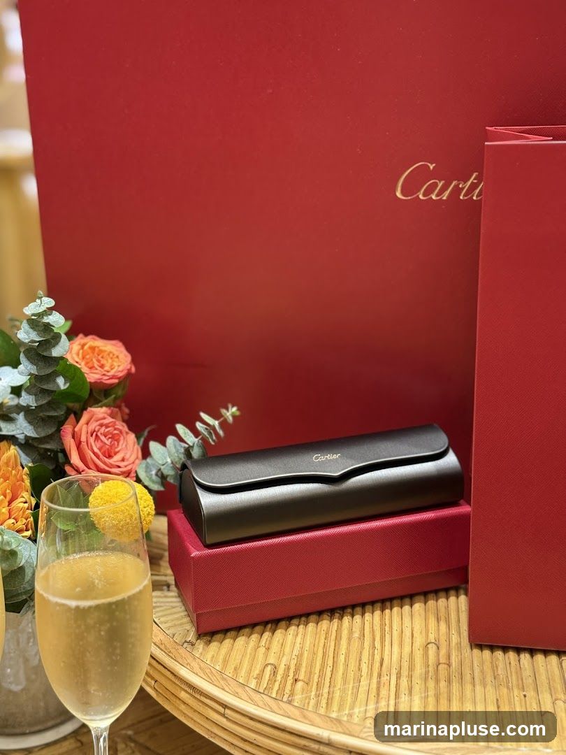 A dark brown Cartier branded eyewear case rests on a red Cartier box. In the foreground, a glass of champagne with a yellow garnish sits next to a bouquet of orange and pink roses and eucalyptus leaves. Two red Cartier boxes are visible in the background.