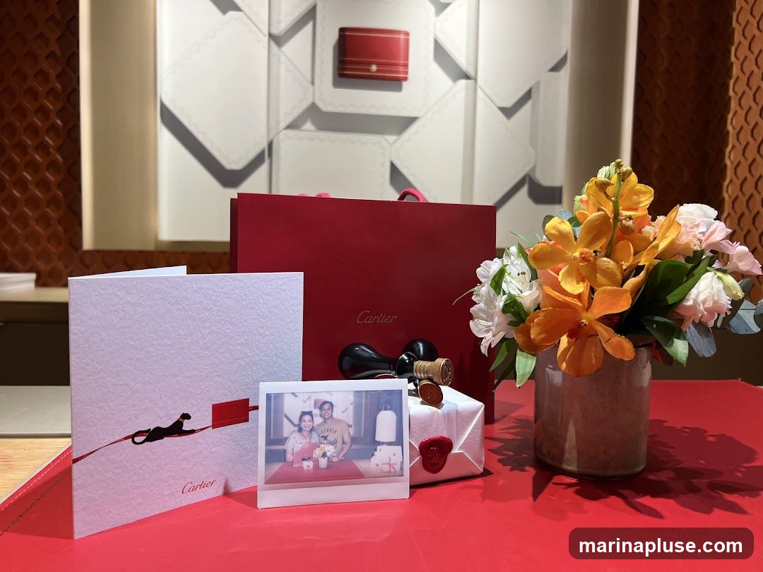 A still life arrangement featuring a red Cartier shopping bag, a white card with a black panther silhouette and the Cartier logo, a small framed polaroid photo of a couple, a wrapped gift with red wax seals, and a vase of orange and white flowers. The background appears to be a display wall with geometric patterns and a textured brown panel.
